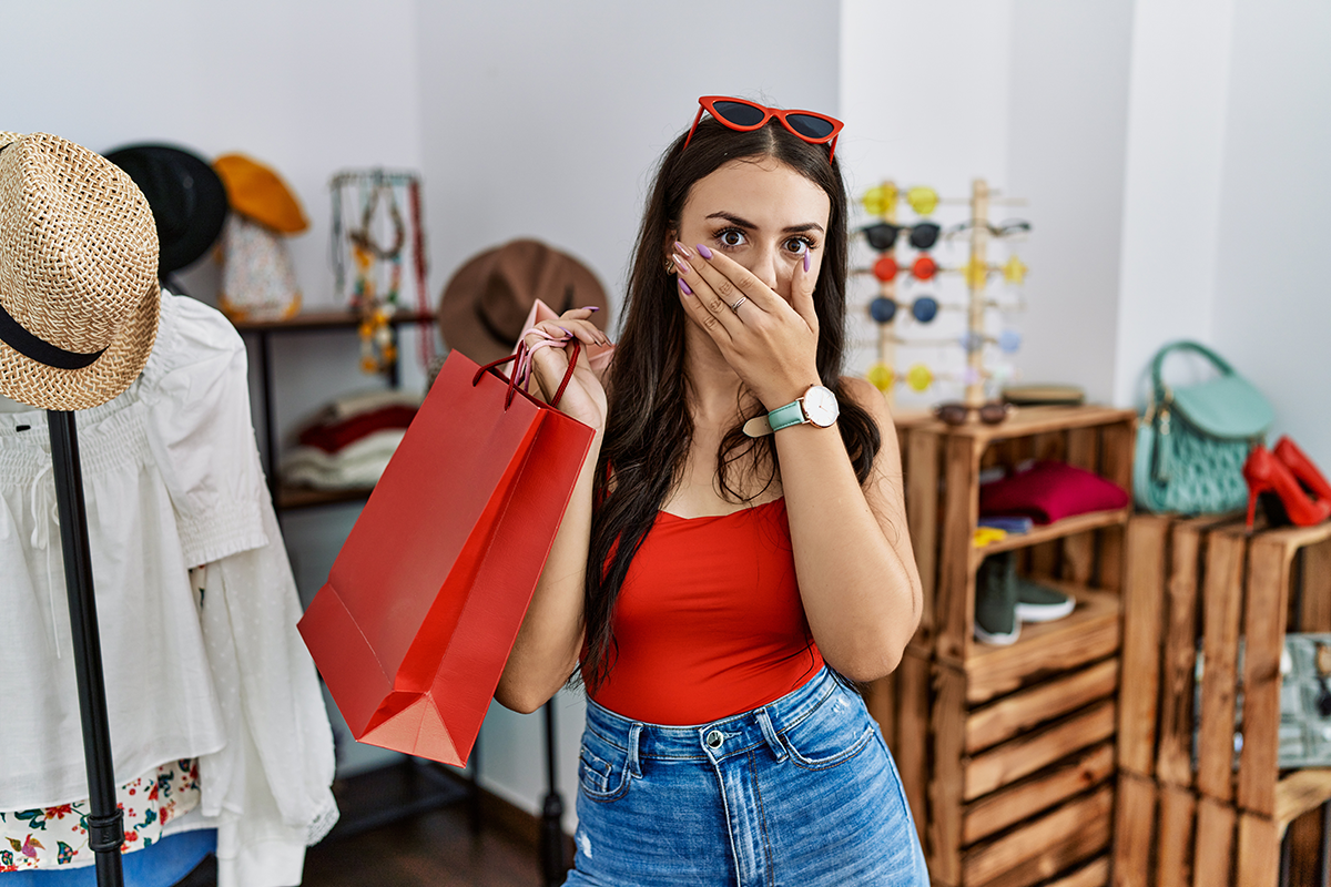 Young brunette woman holding shopping bags at retail shop shocked covering mouth with hands for mistake