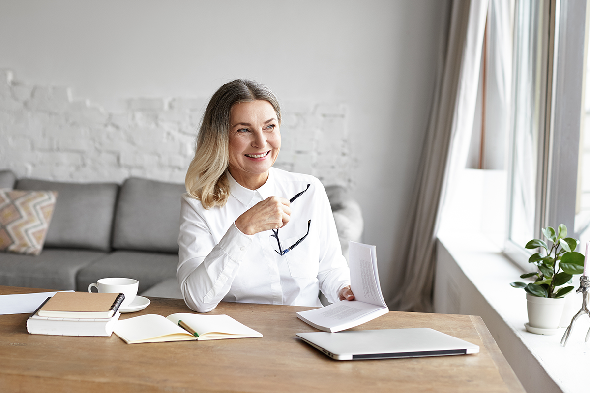 Cheerful middle aged woman smiling broadly, enjoying reading good book during small break while working distantly from home