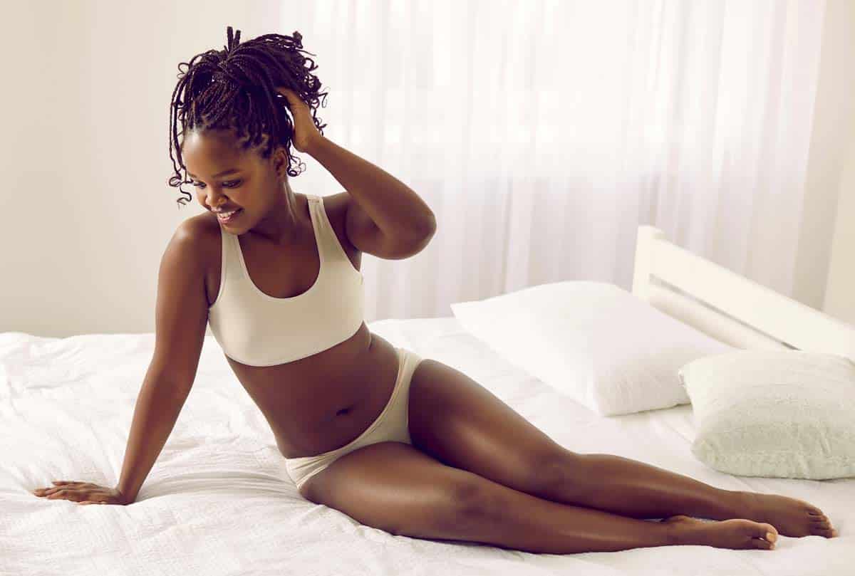 Happy beautiful chubby young African American woman in underwear relaxing on bed at home.